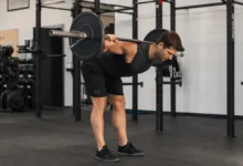 Fit man performing a barbell good morning exercise with the barbell resting across his upper back, hinging forward at the hips while maintaining a neutral spine in a modern gym