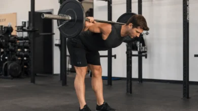 Fit man performing a barbell good morning exercise with the barbell resting across his upper back, hinging forward at the hips while maintaining a neutral spine in a modern gym