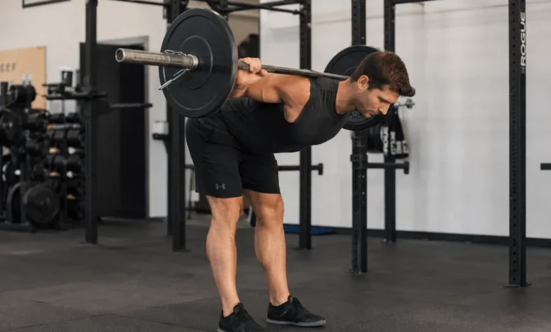 Fit man performing a barbell good morning exercise with the barbell resting across his upper back, hinging forward at the hips while maintaining a neutral spine in a modern gym